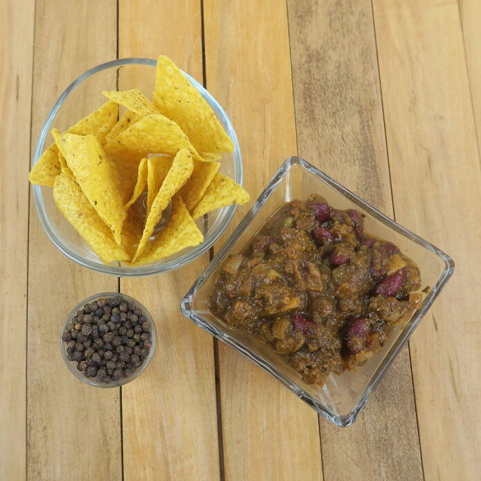 Three glass vessels on a wooden background. A small one contains peppercorns. A larger round one contains yellow corn chips. A square container has Two Alarm No Nightshade Chili, so ground beef and kidney beans in a red sauce.