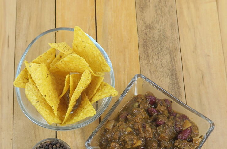 Three glass vessels on a wooden background. A small one contains peppercorns. A larger round one contains yellow corn chips. A square container has Two Alarm No Nightshade Chili, so ground beef and kidney beans in a red sauce.